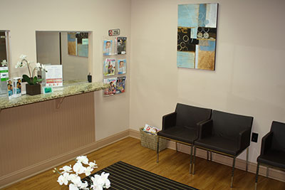 The image shows an interior view of a dental office reception area, featuring a counter with a sink, a waiting area with chairs and a table, a receptionist s desk with a computer monitor, and a wall-mounted framed picture.
