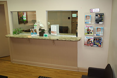 The image is a photograph of an interior space that appears to be a reception area or waiting room, featuring a counter with a glass top and a sign indicating it s for  Reception  and  Check-in,  with pamphlets arranged on the counter. A wall-mounted magazine rack displays various magazines, and there is a potted plant on the counter. The setting suggests a professional environment, possibly within a medical or dental office.