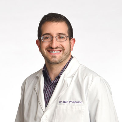 The image features a man wearing glasses, smiling at the camera with his head slightly tilted. He is dressed in a white lab coat and has a beard. Behind him is a plain background.