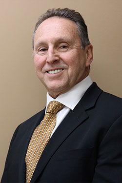 The image shows a man standing in front of a beige background. He is wearing a dark suit and has short gray hair. His expression is neutral, and he appears to be posing for the photograph.