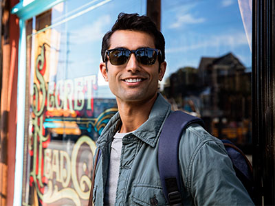 A man in sunglasses, standing in front of a store window, posing with a backpack and smiling.