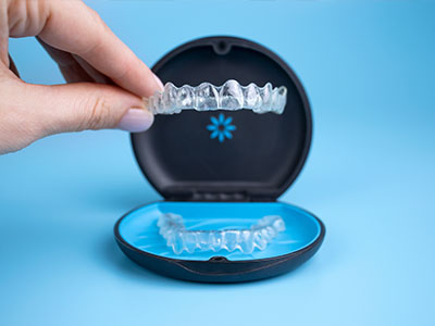 The image shows a person s hand holding an open blue and black dental implant case with a clear plastic tray containing what appears to be a set of artificial teeth, which are placed in front of the case.