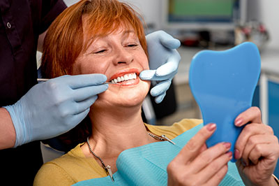 An elderly woman with a toothy smile is seated in a dental chair, receiving a teeth cleaning or dental treatment from a dental professional.