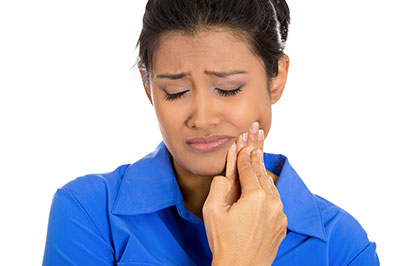 A woman with dark hair, wearing a blue shirt, holds her hand to her face and appears to be in distress or discomfort.