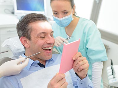 The image shows a man sitting in a dental chair, holding a pink card with a surprised expression on his face, while a dentist and a hygienist look at him.
