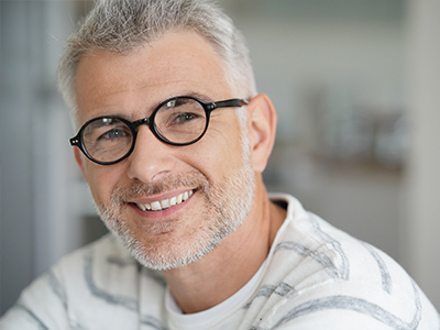 The image shows a man with gray hair, wearing glasses and a white shirt, smiling at the camera.