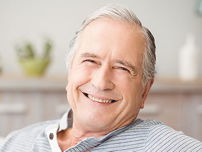 The image shows an elderly man with a smile, sitting in a chair and looking directly at the camera.