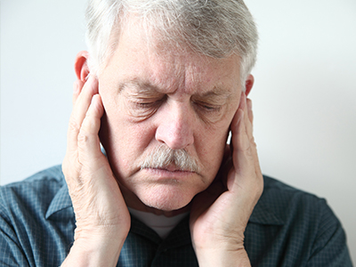 The image shows an older man with a beard, wearing glasses and holding his hand to his ear. He appears to be in deep thought or possibly experiencing discomfort.